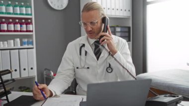 Modern klinikte doktor telefonla konuşuyor. Stetoskop ve beyaz önlüklü. Etrafı dosyalar ve ilaç şişeleriyle çevrili..