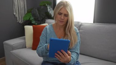 Tablet ve kredi kartını şık dekorlu, mavi süveter ve kot pantolon giymiş, rahat oturma odasında online alışveriş yapmak için kullanan bir kadın..