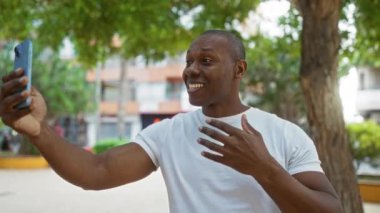 Genç adam, şehir parkında ağaçlar ve binalar eşliğinde akıllı telefondan video sohbeti yaparken gülümsüyor.