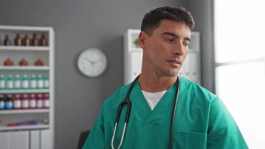 Hastane odasında steteskoplu yeşil önlük giyen genç adam kendinden emin ve profesyonel görünüyor.