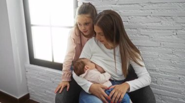 Anne bebeğini emzirirken küçük kız sevgi dolu bir şekilde yatak odası ortamını seyrediyor, aile sıcaklığını ve bağlantısını gösteriyor..