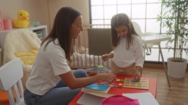 Kadın ve çocuk rahat bir çocuk odasında eğitici oyuncaklarla oynuyor, kırmızı bir masada oyuncaklarla öğrenme ve kaynaşma için sıcak ve etkileşimli bir ortam yaratıyorlar..