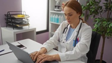 Klinikte dizüstü bilgisayarda çalışan, beyaz önlüklü, boynunda steteskop ve arkasında tıbbi malzemeler olan bir kadın..
