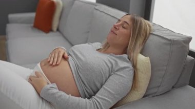 Hamile bir kadın, rahat bir oturma odasında, rahat bir oturma odasında, elini karnına koyup, rahat bir evde anne beklediği huzurlu bir anı yakalıyor..