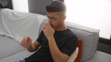 İçerideki rahat bir kanepede dikkatlice oturan genç adam, gözlüklü, siyah gömlekli, rahat bir ev ortamında düşünceli bir yüz ifadesi sergiliyor..