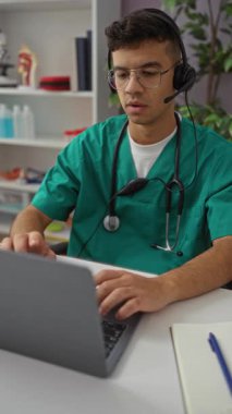 Klinik ortamda kulaklıkları çıkarırken stresli görünen genç bir adam, kapalı bir hastane ortamında dizüstü bilgisayarın yanında yeşil önlükler giyiyor..