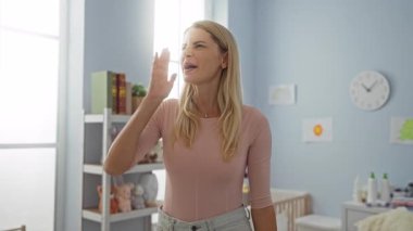 Bebek odasında bilgisiz bir kadın, hiçbir fikri olmayan genç bir sarışın, kuşkular içinde kollarını kaldırmış ve kafası karışmış.