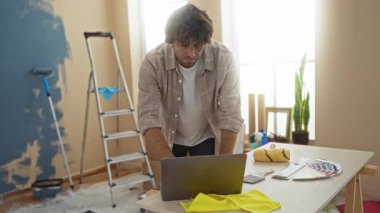 Oturma odasında duvarları maviye boyayan genç bir adam, yakınlarda fırça ve merdiven bulunan bir masada dizüstü bilgisayarı kontrol ederken, ev yenileme ve yaratıcılığı somutlaştırıyor..