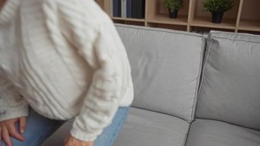 Oturma odasında rahat bir kanepede dinlenen beyaz bir kazak ve arka planda kitaplığı olan mavi bir kot pantolon giyen bir kadın rahat bir ev atmosferini işaret ediyor..