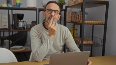 Orta yaşlı İspanyol bir adam modern iç mekan ve mutfak malzemeleriyle çevrili dizüstü bilgisayarını kullanırken fikir yürütüyor..