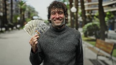 Gülümseyen İspanyol adam, Amerikan dolarlarını neşeyle elinde tutuyor şehir sokaklarında başarıyı ve finansmanı sergiliyor..