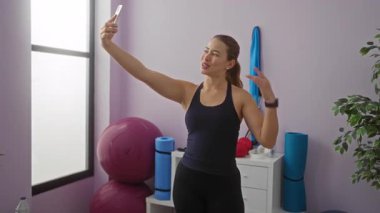 Spor salonunda elinde telefonla gülümseyen bir kadın spor kıyafetleri giyip spor aletleri ve bitkilerle telefonda konuşuyor..