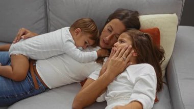 Kadın, oğlu ve kızıyla birlikte rahat bir oturma odasında, evde sevgi ve birlikteliği resmediyor..