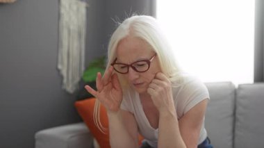 Oturma odasında albinizmi olan bir kadın gözlüğünü çıkarıp gözlerini ovalıyor. Ev ortamında yorgun ve düşünceli görünüyor..
