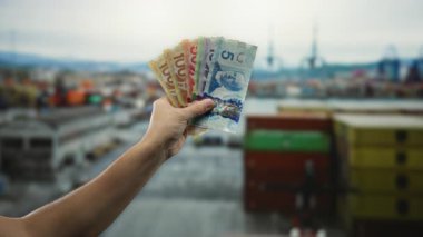 Deniz kıyısındaki limanda su ve teknelerle çevrili Kanada banknotları tutan adam endüstriyel arka plana karşı canlı para birimine odaklanıyor..