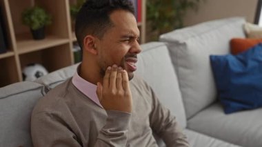 Diş ağrısı çeken genç bir adam, rahat bir oturma odasında rahat bir koltukta oturuyor. Yüzündeki rahatsızlık ifadesi, yumuşak yastıklar ve iç dekorlarla çevrili..