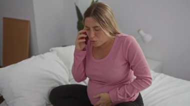 Hamile kadın kucağında karnıyla yatakta oturuyor ve rahat yatak odasında telefonla konuşuyor. Rahat bir dekorla çevrili yaklaşan bebek için karışık heyecan ve endişe duygularını dile getiriyor..