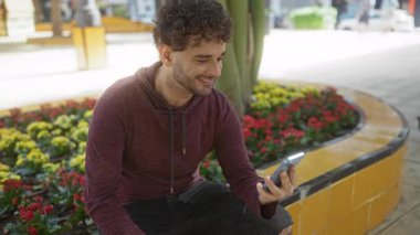 Şehir parkında oturan genç adam canlı çiçekler ve şehir hayatı ile çevrili telefonuna bakarken gülümsüyor..