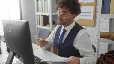 Ofiste çalışan genç bir adam bilgisayar ve belgelerle çalışıyor, gözlük ve yelek takıyor, odaklanmış bir iş ortamını gösteriyor..