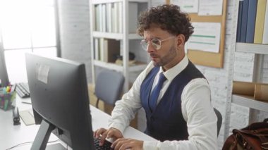 Modern ofis ortamında bilgisayar üzerinde yoğun çalışan İspanyol bir adam, çağdaş çalışma alanındaki kitaplıklar ve ofis malzemeleriyle çevrili, odaklanma ve profesyonellik sergiliyor..