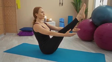 Spor salonunda yoga yapan bir kadın, evin içinde renkli spor ekipmanlarıyla çevrili bir paspasın üzerinde pilates yapıyor..