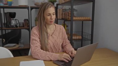 Modern ofis ortamında dizüstü bilgisayarda çalışan, koluna hapşıran, pembe kazak giyen, raflı, arka planda mutfak aletleri olan, günlük iş hayatını gösteren bir kadın..