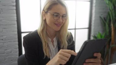 Sarı saçlı ve gözlüklü bir kadın modern ofis ortamında büyük pencereli ve bitkilerli bir tablet kullanıyor.