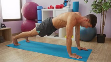 Spor salonunda mavi bir paspasın üzerinde egzersiz yapan genç bir adam el yaralanmasının acısını gösteriyor. Spor aletleri ve yoga toplarıyla çevrili..