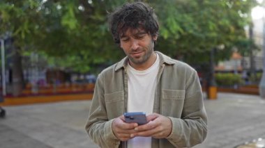 Genç adam, arka planda bulanık ağaçlar ve insanların bulunduğu bir şehir parkında akıllı telefon kullanırken gülümsüyor.