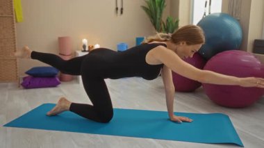 Spor salonundaki mavi minderde yoga yapan bir kadın. Dengeli toplar ve mumlarla dingin bir atmosfer yaratıyor..