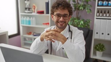 Klinikteki genç İspanyol erkek doktor, arka planda dizüstü bilgisayarı ve tıbbi ekipmanlarla bir masada otururken kendinden emin bir şekilde gülümsüyor..