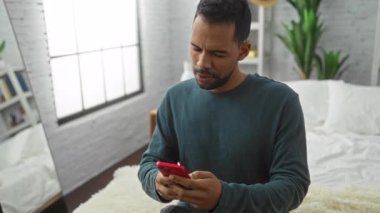 Modern yatak odasında akıllı telefon kullanan genç adam yatağın kenarında düşünceli bir anı yakalıyor..