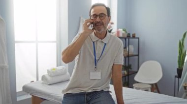 Rehabilitasyon kliniğindeki İspanyol adam telefonla konuşuyor, gözlük takıyor ve günlük giysiler giyiyor, en az dekorla aydınlık odada terapi masasında oturuyor..