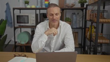 Beyaz gömlekli bir adam masanın üzerinde gözlüklü bir şekilde oturmuş modern ofiste dizüstü bilgisayara bakıyor. Etrafı rahat bir ortamda raflar ve bitkilerle çevrili.