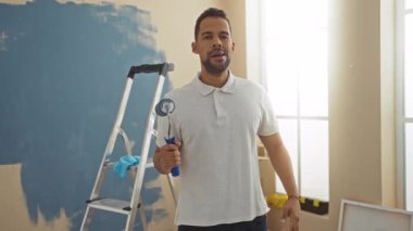 İspanyol adam oturma odasında paten ve merdivenle resim yaparken jest yapıyor. İçerde eğlenceli bir yenileme projesi öneriyor.