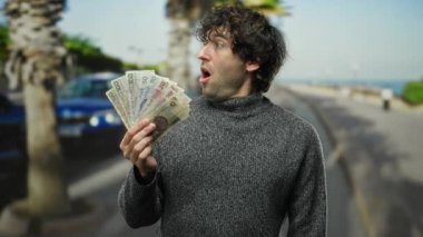 İspanyol adam şehir caddesinde elinde Czech Koruna banknotlarıyla dikiliyor, şaşırmış bakıyor, ağaçlar ve arabalarla çevrili..