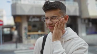 Gözlüklü genç adam şehir sokaklarında diş ağrısı çekiyor, beyaz bir kapüşonlu giyiyor, rahatsızlığını dile getiriyor, şehir geçmişi var, ortak diş sorunlarını vurguluyor..