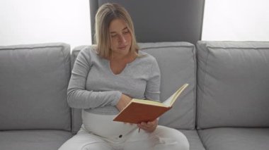 Hamile kadın, rahat oturma odasının iç tarafındaki kanepede kitap okuyor. Huzurlu ve samimi ev atmosferini ifade ediyor..