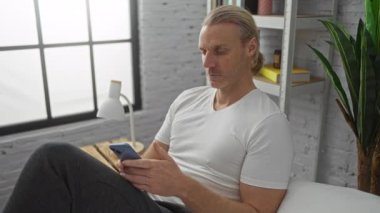 Orta yaşlı, beyaz bir adam akıllı telefonuyla konuşuyor. Parlak, modern yatak odasında yatakta oturuyor..