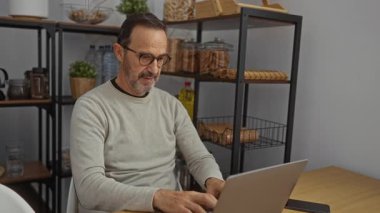 Samimi bir ofiste dizüstü bilgisayarda çalışan, çeşitli eşyalarla dolu rafları olan olgun İspanyol bir adam, odaklanmış bir çalışma alanı oluşturur..