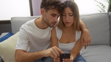 Çift, rahat bir oturma odasında akıllı telefon kullanarak kanepede oturuyor ve bir kadın ve erkeğin teknolojiyle iç içe olduğu bir anı öneriyor.