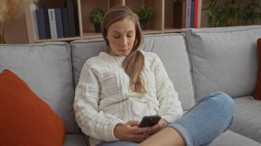 Modern oturma odasındaki kanepede oturan kadın rahat bir ev dekoru ve arka planda kitaplıkla çevrili günlük beyaz bir kazak ve kot pantolon giyen düşünceli bir ifadeyle telefona bakıyor..