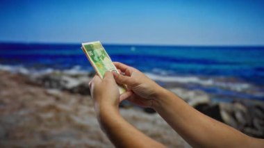 El ele tutuşan İsrail şekeli banknotlar manzaralı bir sahil arka planına karşı, açık deniz kenarında finans ve yatırım kavramlarını tasvir ediyor.