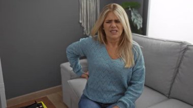 Rahat bir oturma odasında gri bir kanepede otururken sırt ağrısını ifade eden bir kadın bir rahatsızlık anı ve ev ortamını yakalıyor..