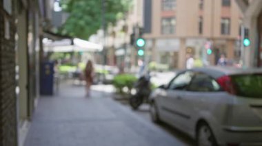 Bulanık bir kadının güneşli havada binaların ve arabaların yanında yürüdüğü bir açık hava manzarası.