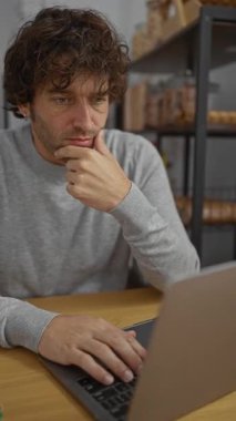 Rafların arka planında dizüstü bilgisayar kullanan düşünceli bir adam, profesyonel bir çalışma alanında odaklanmayı ve düşünmeyi vurguladı..