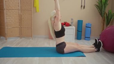 Kadın yoga minderi ve ağırlıklarla spor yapıyor. Parlak odada spor aletleri ile çevrili. Güç ve kararlılık gösteriyor.