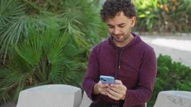 Genç adam, yeşil yemyeşil yeşilliklerle çevrili güneşli şehir parkında akıllı telefon kullanırken gülümsüyor..