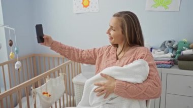 Kadın pembe kazaklı bebekle birlikte gülümsüyor rahat bir yatak odasında oturmuş modern evdeki neşeli aile anlarını gösteriyor..