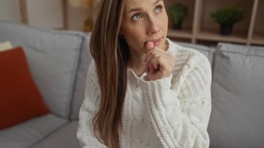 Tırnaklarını yiyen çekici genç bir kadın, rahat bir oturma odasında, beyaz bir kazak giyip, bir apartmanın içindeki rahat bir koltuğa oturmuş, düşünceli görünüyor..